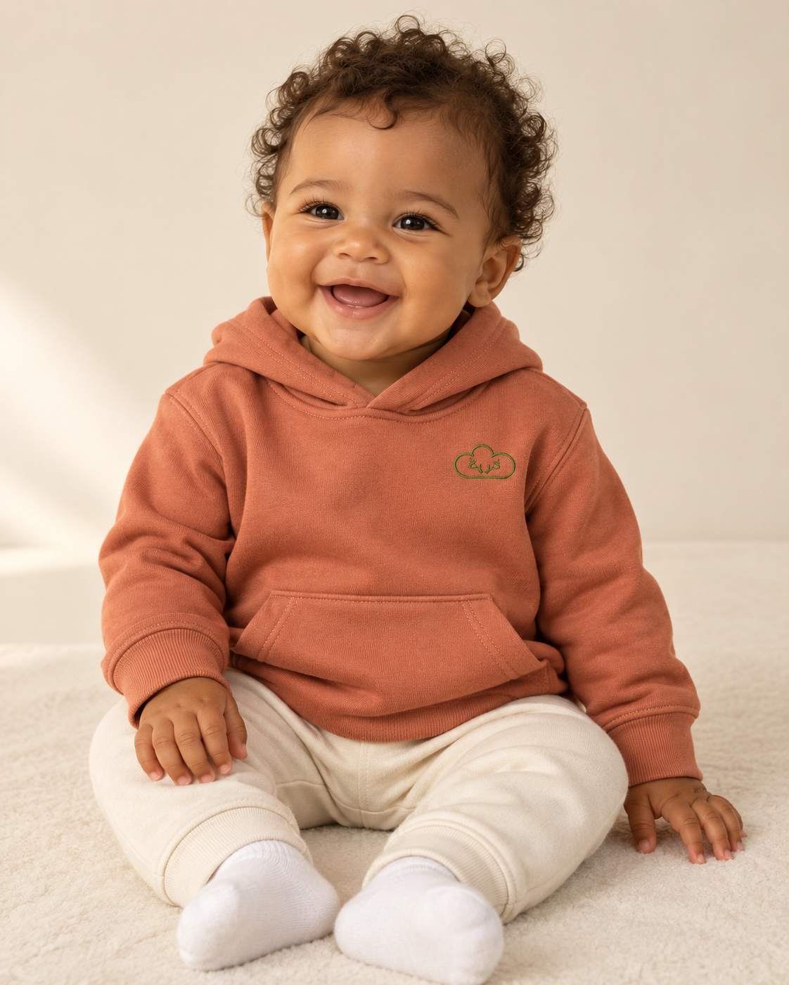 Baby wearing a terracotta Hunted Hoodie with a green cloud-and-antler chest emblem.