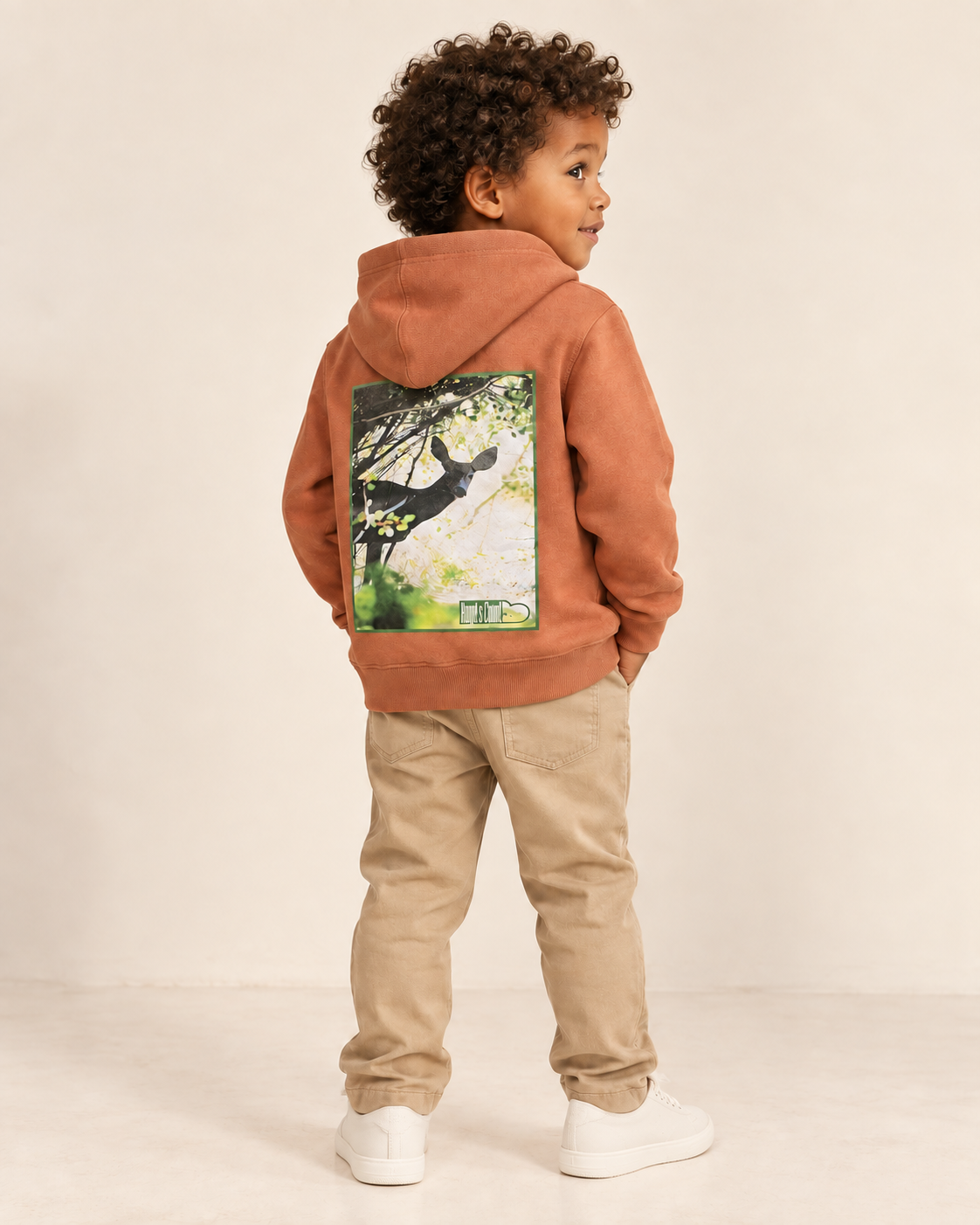 Child wearing a terracotta Hunted Hoodie, with the green-framed deer print visible on the back.